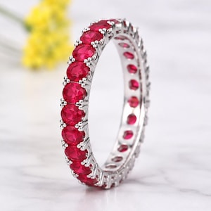 May include: A silver eternity band featuring a continuous row of round, vibrant red gemstones set in a polished metal frame. The ring is positioned upright, showcasing the uniform circular stones that encircle the entire band in a classic design.