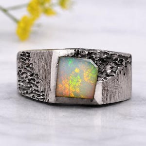 Op de afbeelding: Een zilveren ring met een vierkant geslepen opaal met iriserende flitsen van groen, oranje en geel. De band heeft een donkere, geoxideerde afwerking met een zwaar getextureerd, ruw oppervlak dat contrasteert met de gladde, gepolijste edelsteen.