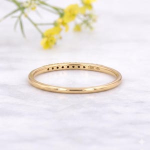 May include: A delicate gold ring, featuring tiny gemstones set along its band. The ring is stamped with "18K" and a hallmark. The background shows a marble surface and yellow flowers. The ring's design is simple and elegant.