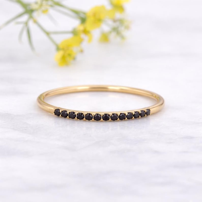 May include: A thin gold band ring featuring a row of small, round, black gemstones set along the front half of the band. The metal has a polished finish, and the stones are uniform in size, creating a continuous line of dark accents.