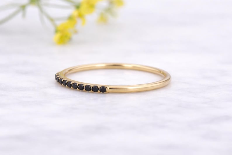 May include: A gold ring featuring a row of small, black gemstones. The ring is set against a white marble surface, with a blurred background of yellow flowers.