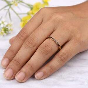 May include: A gold ring featuring a line of small, black gemstones. The ring is on a finger, with the hand in the foreground. The background is blurred, with suggestions of yellow and white flowers.