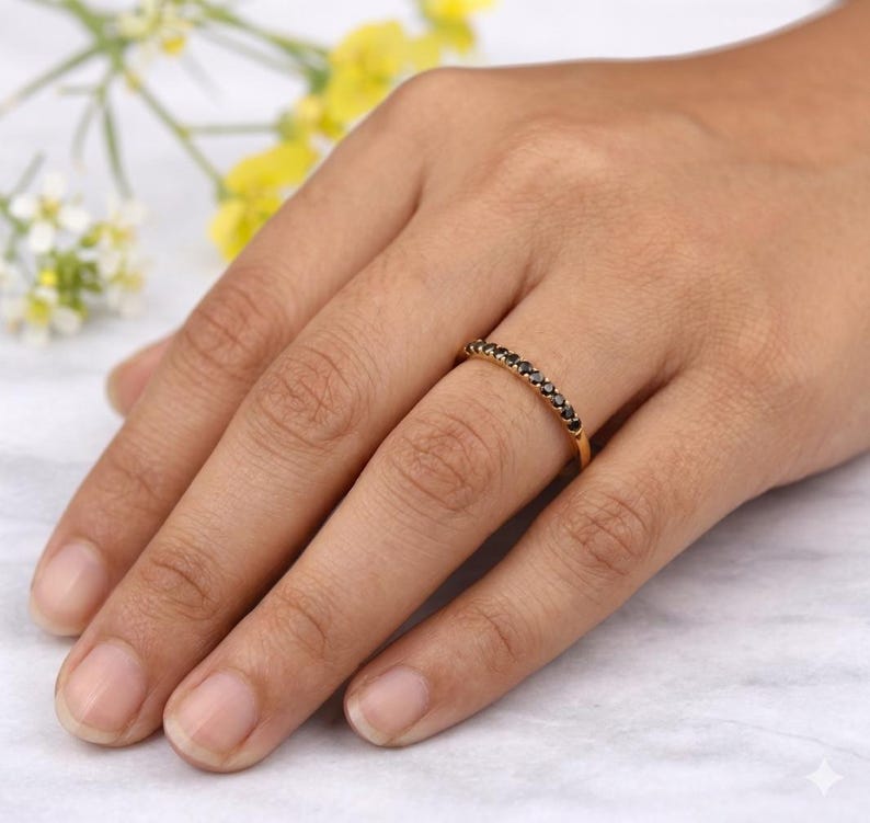 May include: A gold ring featuring a line of small, black gemstones. The ring is on a finger, with the hand in the foreground. The background is blurred, with suggestions of yellow and white flowers.