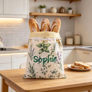 May include: A cream-colored fabric bread bag featuring a botanical print of green herbs and purple lavender sprigs. The name Sophie is embroidered in dark green script on the front. Three crusty baguettes are tucked inside the open top.