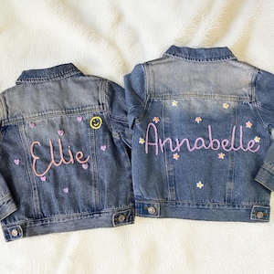 May include: Two denim jackets with personalized embroidery. One jacket features the name "Ellie" in pink script with heart and smiley face accents. The other jacket has the name "Annabelle" in pink script with flower accents.