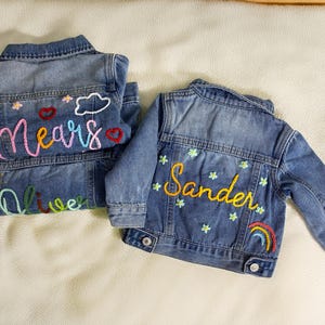 May include: Two blue denim jackets with embroidered names and designs. One says "Mears" with hearts, a cloud, and a sun. The other says "Sander" with flowers and a rainbow.