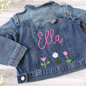 May include: A blue denim jacket with embroidered details. The back features the name "Ella" in pink script, a bee, and floral designs. The jacket has button closures and long sleeves.