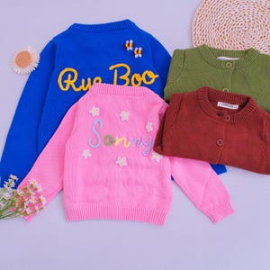 Könnte beinhalten: Eine Auswahl von vier langärmeligen Pullovern in Blau, Pink, Olivgrün und Kastanienbraun. Der blaue Pullover zeigt den gelben Sticktext Rue Boo und zwei kleine Bienen. Der pinke Pullover trägt den gestickten Namen Sonny und weiße Blumen.