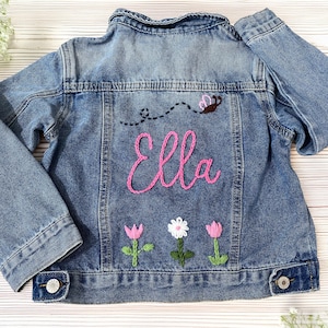 May include: A blue denim jacket with embroidered details. The back features the name "Ella" in pink script, a bee, and three embroidered flowers. The jacket has button closures and pockets, suitable for children's apparel.