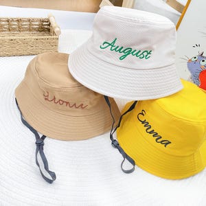 Könnte beinhalten: Drei Fischerhüte in Beige, Weiß und Gelb. Der weiße Hut hat "August" in Grün gestickt. Der beige Hut hat "Lioni" in einer passenden Farbe gestickt. Der gelbe Hut hat "Emma" in Schwarz gestickt. Jeder Hut hat einen Kinnriemen.