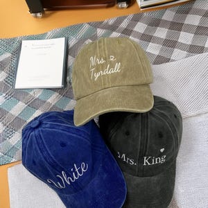 Op de afbeelding: Drie baseballpetten in olijfgroen, marineblauw en zwart. Op de olijfgroene pet staat "Mrs. Tyndall" geborduurd. Op de marineblauwe pet staat "White" geborduurd. Op de zwarte pet staat "Mrs. King" geborduurd.