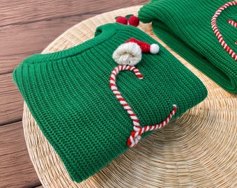 May include: A vibrant green knit sweater featuring a three-dimensional candy cane design made of red and white yarn. The sweater is adorned with a small white and red Santa hat applique and two small red pom-poms near the ribbed crew neckline.