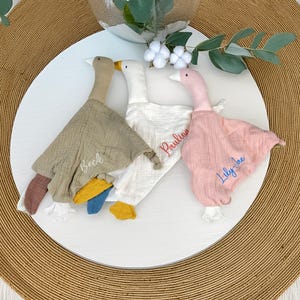 May include: Three goose-shaped baby comforters in beige, white, and pink, each with a personalized name embroidered on the fabric. The comforters have colorful accents on the feet and wings. The names embroidered are "Beck", "Paulina", and "Lily-Jae".