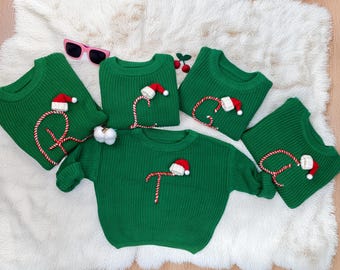 May include: Five green knit sweaters, each featuring a letter formed by a red and white striped candy cane pattern topped with a small red and white Santa hat. The letters displayed are R, L, E, A, and T. A pair of pink sunglasses is placed near the top.