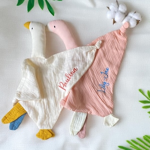 May include: Two goose-shaped baby comfort blankets. One is white with yellow and blue accents, the other is pink. Each has a knotted head and a name embroidered on the blanket: "Paulina" and "Lily-Jae". Cotton buds are attached to one blanket.