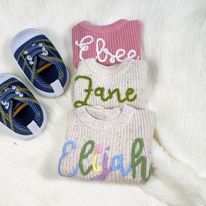 May include: Three personalised baby sweaters in pink and speckled cream, each embroidered with a name in a different colour. A pair of blue baby shoes with yellow laces are next to the sweaters. The sweaters are made of a soft, knitted material.