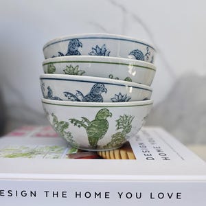 May include: Four white ceramic bowls with a bird and floral pattern in blue and green are stacked. The bowls are on a book with the text "DESIGN THE HOME YOU LOVE" and "HABITAT".