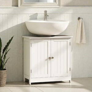 May include: A white bathroom vanity unit with a vessel sink. The unit has two doors with vertical paneling and round gold-coloured knobs. A chrome tap is mounted on the sink. A white towel hangs on a nearby towel rail. A potted plant is on the left.