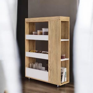May include: A light wood rolling storage cart with white shelves and drawers. The cart has multiple shelves and drawers, and is filled with various items, including jars, glasses, and books. The cart is on wheels for easy movement.