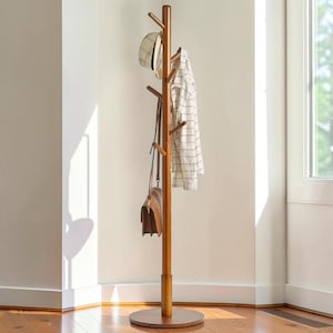 Puede incluir: Un perchero alto de madera marrón con múltiples ramas para colgar objetos. Un sombrero, un bolso y una camisa a cuadros cuelgan del perchero. El fondo es una pared clara y una ventana.
