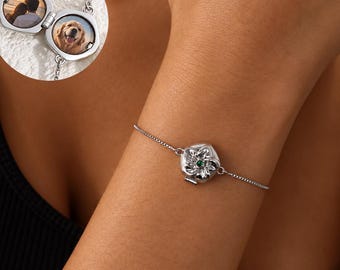 May include: A silver-toned locket bracelet featuring a hexagonal charm with a small green stone at the center. The locket opens to reveal two circular photo frames, one containing a picture of a person and the other a golden retriever dog.