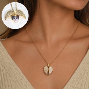 Può includere: Una collana color oro con un ciondolo a forma di cuore dotato di due ali d'angelo incernierate e incastonate di cristalli, che si aprono per rivelare una piccola foto a forma di cuore di un gatto. Il ciondolo pende da una sottile catenina.