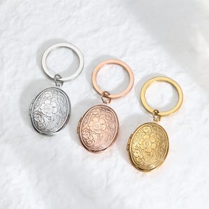 May include: Three oval-shaped locket keychains in silver, rose gold, and gold. Each locket features a floral design and is attached to a round keyring. The lockets are displayed on a white, textured surface.