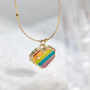 May include: A gold-colored necklace with a heart-shaped pendant. The pendant features a rainbow-colored striped design with red, orange, yellow, green, blue, and purple stripes. The necklace has a delicate chain.