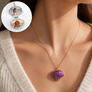 May include: A gold-colored necklace with a purple seashell pendant. The necklace has a delicate chain and a seashell-shaped pendant with a gold-colored outline. The pendant is purple with gold-colored accents. A silver locket with two photos is also visible.