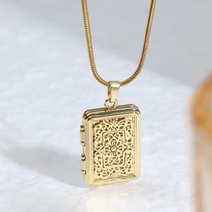 May include: A gold-colored rectangular pendant necklace with an ornate floral design. The pendant is suspended from a delicate gold chain. The pendant has a hinged design, suggesting it may open. The background is a soft, neutral color.