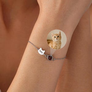 May include: A silver-toned chain bracelet featuring a small cat-ear shaped charm engraved with the name Bailey and a circular pendant containing a projection of a ginger kitten. The delicate metal links connect these two distinct focal elements.