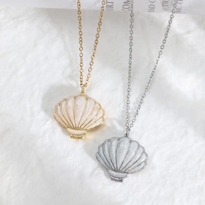 Può includere: Due collane con ciondoli a forma di conchiglia. Un ciondolo è color oro con una finitura scintillante bianco sporco, l'altro è color argento con una finitura scintillante bianca. Entrambi i ciondoli pendono da sottili catene a maglie metalliche.