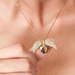 Könnte beinhalten: Ein goldfarbener herzförmiger Medaillon-Anhänger an einer feinen Kette. Das Medaillon verfügt über zwei bewegliche Engelsflügel, die mit klaren Kristallen besetzt sind. In der Mitte des Herzens befindet sich ein kleines, rundes Foto einer älteren Person.