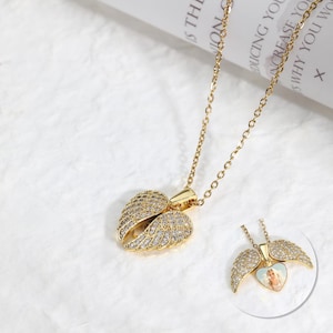 May include: Gold-colored necklace with a pendant shaped like angel wings. The wings are encrusted with small, clear crystals. A smaller version of the necklace is visible, showing a heart-shaped photo inside the pendant.