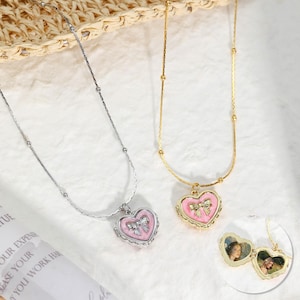 May include: Two heart-shaped locket necklaces. One is silver with a pink heart and bow pendant, the other is gold with a pink heart and bow pendant. The gold necklace has a small photo inside the locket.