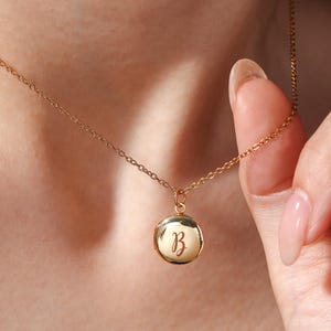May include: A gold-colored necklace with a round pendant. The pendant features the letter "B" in a cursive script. The necklace is worn around the neck, and the chain is visible. The pendant is approximately 0.75 inches in diameter.