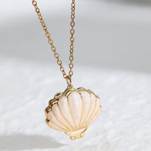 May include: A gold necklace with a seashell pendant. The pendant is white with gold edging and hangs from a fine chain. The seashell design is detailed and has a hinged opening.