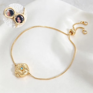 Könnte beinhalten: Ein goldfarbenes Medaillon-Armband mit einem blütenförmigen Anhänger und einem kleinen blauen Stein in der Mitte. Das Medaillon lässt sich öffnen und zeigt zwei runde Fotoeinsätze. Das Armband hat eine feine Kette und einen verstellbaren Verschluss.