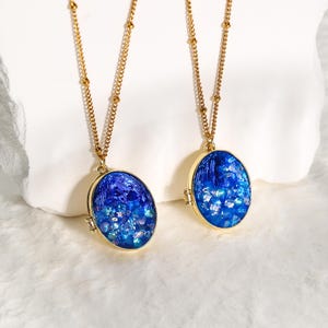 Puede incluir: Dos collares dorados con colgantes ovalados. Los colgantes tienen un diseño azul brillante dentro de un marco dorado. Las delicadas cadenas tienen pequeñas cuentas redondas. El fondo es una superficie blanca y suave.