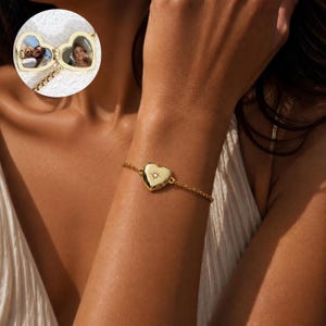 May include: A gold-toned heart-shaped locket bracelet featuring a central star-engraved accent with a small embedded stone. The locket opens to reveal two circular photo frames containing images of individuals, attached to a delicate gold-toned chain.
