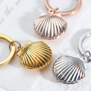 Pode incluir: Close-up de três chaveiros em forma de concha em ouro, ouro rosa e prata. Cada chaveiro tem um anel circular e um pingente de concha detalhado e canelado. Os chaveiros são exibidos em uma superfície branca texturizada.