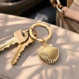 May include: A gold-toned metal keychain featuring a textured seashell charm attached to a circular split ring. Two standard brass-colored keys are also attached to the ring, which has a polished finish and a distinct ribbed pattern on the shell.