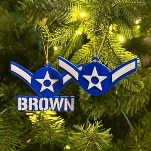 May include: Two blue and white Christmas ornaments with a star and wing design, and the name "BROWN" in white block letters. The ornaments are hanging on a green Christmas tree.