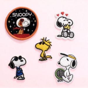 Puede incluir: Parches bordados con los personajes de Snoopy y Woodstock. Un parche muestra a Snoopy con un traje espacial con la palabra "SNOOPY". Otros parches representan a Snoopy con corazones, gafas de sol y una raqueta de tenis. Los parches están sobre un fondo rosa.