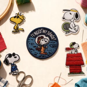 May include: Embroidered patches featuring Snoopy and Woodstock characters in various poses. One patch reads "I NEED MY SPACE." Other patches show Snoopy playing tennis, wearing a pilot helmet, and on top of his doghouse. Sewing supplies are also visible.