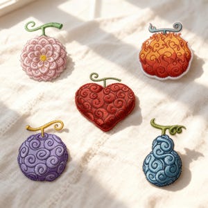 Puede incluir: Parches bordados con diseños de frutas variadas, incluyendo una flor rosa, un corazón, una forma de llama, una esfera morada y una pera azul. Cada parche presenta costuras detalladas y un tallo. Los parches están dispuestos sobre una superficie blanca texturizada.