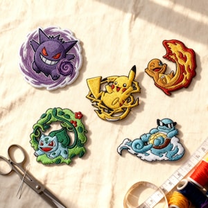 Op de afbeelding: Geborduurde patches met verschillende stripfiguren in levendige kleuren. De patches tonen een paarse Gengar, een gele Pikachu, een oranje Charmander, een groene Bulbasaur en een blauwe Squirtle. Naaibenodigdheden zijn zichtbaar op de achtergrond.