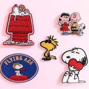 May include: A selection of embroidered Peanuts character patches. The patches depict Snoopy in his doghouse, Woodstock, Snoopy with a heart, and other characters. The patches are in various colours, including red, blue, yellow, and white.