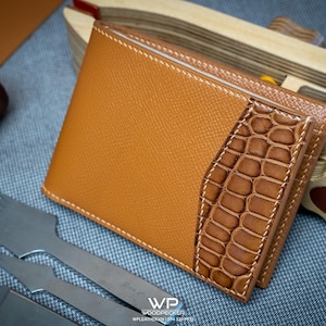 Peut inclure: Un portefeuille en cuir marron clair à deux volets avec une finition texturée. Il présente un panneau contrastant à texture crocodile et des coutures blanches. Des outils et du fil sont visibles, suggérant un article fait à la main. Un accessoire élégant.