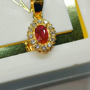 May include: A gold pendant featuring an oval, red gemstone surrounded by small, clear gemstones. The pendant is set in a gold frame and hangs from a gold chain. The jewelry is displayed in a white box.